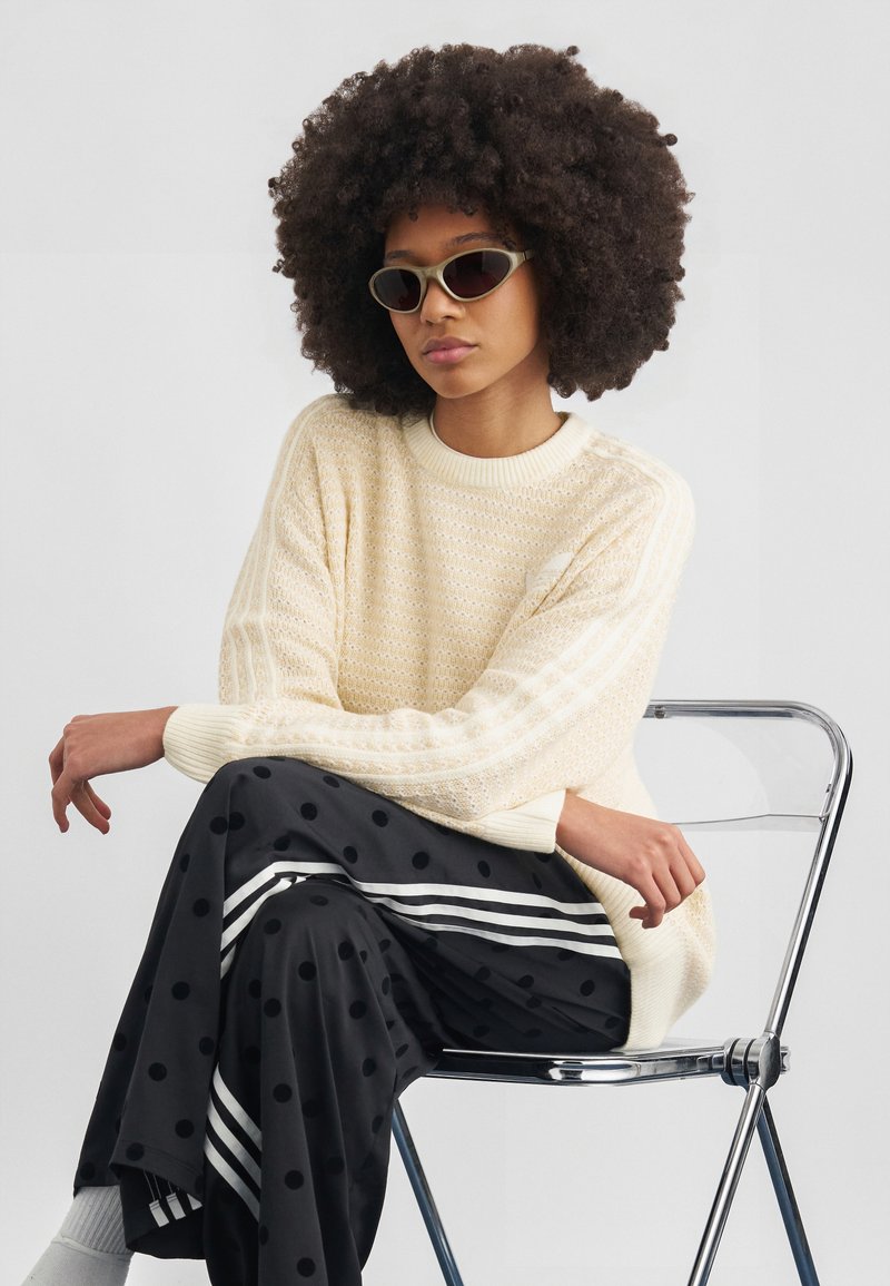 Γυναίκα με πυκνά σγουρά μαλλιά που κάθεται σε διαφανή καρέκλα, φοράει μπεζ πουλόβερ, μαύρο παντελόνι με πουά και λευκές ρίγες, και γυαλιά ηλίου.