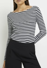 Kvinna i en svart och vit horisontellt randig långärmad topp instoppad i högmidjade svarta byxor mot en vit bakgrund.