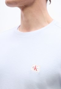 Άτομο που φοράει λευκό που κάμισο με υφή, με μικρό τετράγωνο ταμπελάκι λογότυπου Calvin Klein σε κόκκινο και μπεζ στο στήθος.