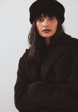 Sieviete ar gariem tumšiem matiem, valkājot melnu pūkainu cepuri un atbilstošu mēteļi, skatās tieši priekšā ar sakrustotām rokām uz balta fona.