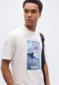 Νεαρός άνδρας με κοντά σγουρά μαλλιά που φοράει ένα λευκό μπλουζάκι με μπλε γραφικό κύμα και το κείμενο «HUGO», κρατώντας μια μαύρη τσάντα με ιμάντα.