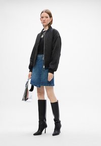 Schwarze Bomberjacke, eng anliegendes schwarzes T-Shirt, jeansfarbener A-Linien-Rock, glänzende silberne Tasche, kniehohe schwarze Stiefel mit Absatz. Einfaches Design in neutralen Farben.