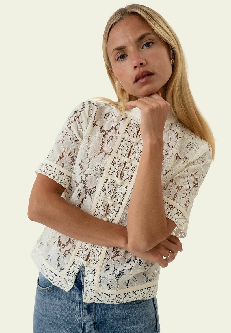 Femme blonde portant un chemisier en dentelle blanche à fleurs avec des manches courtes et un jean bleu, reposant son menton sur sa main.