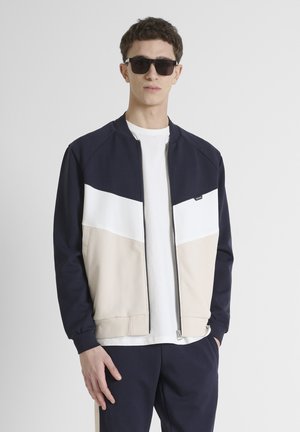 Giacca a zip blu navy e panna con un pannello bianco, realizzata in tessuto morbido. Presenta un colletto alto e tasche, indossata con una t-shirt bianca.