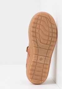 Suela de zapato marrón texturizada con un patrón ondulado. Presenta material de goma con líneas de agarre y un emblema redondo en el centro.