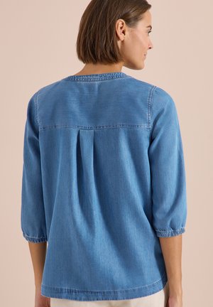 Femme portant un chemisier bleu en denim ample avec des manches trois-quarts et des poignets plissés, vue de dos sur un fond uni.