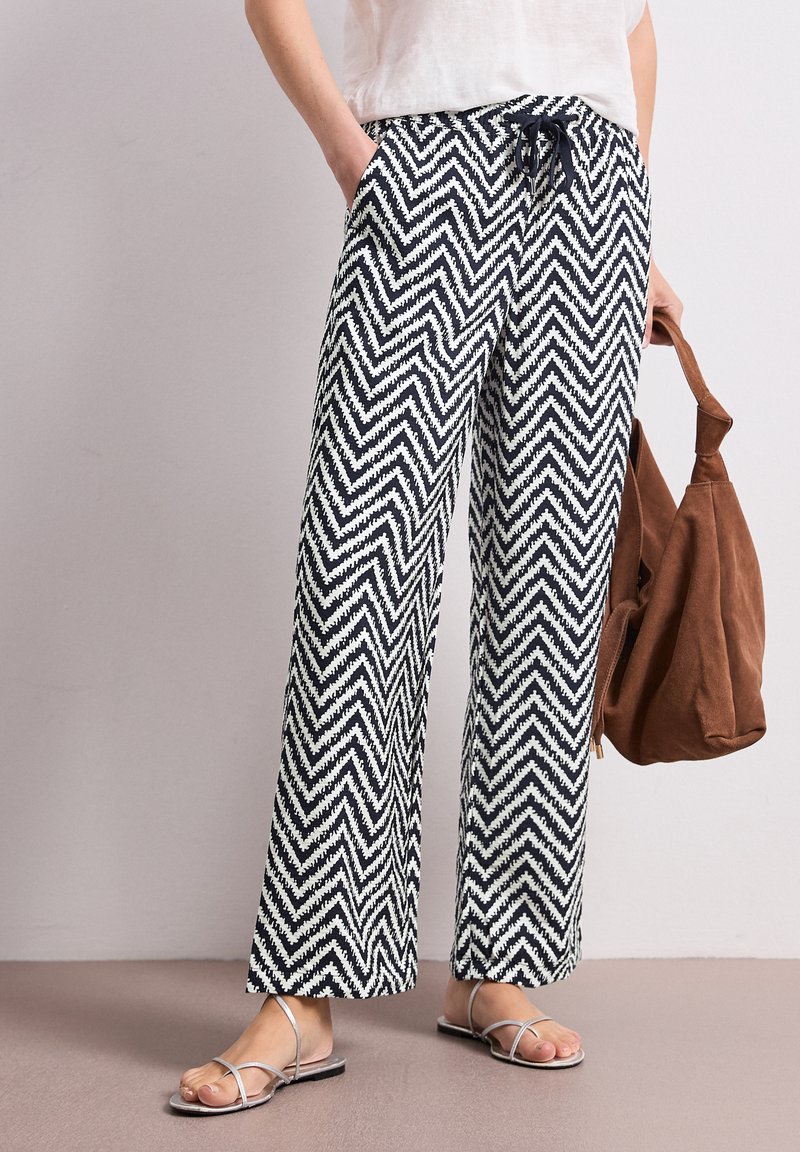 Personne portant un pantalon large à motif zigzag noir et blanc, un haut blanc, des sandales argentées, tenant un sac à main en daim marron.