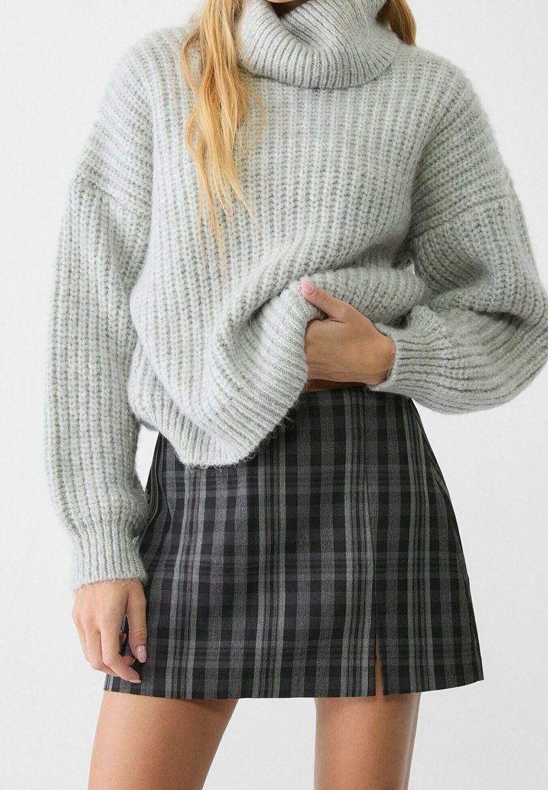 Femme portant un pull à col roulé en maille épaisse grise et une mini-jupe à carreaux noire et grise avec une fente à l'avant.