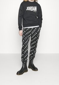 Felpa nera a maniche lunghe con la scritta "JORDAN" in bianco, abbinata a pantaloni jogger neri con il logo "JORDAN" bianco ripetuto e stivaletti neri alla caviglia.