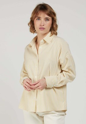 Femme aux cheveux courts et ondulés portant une chemise ample à rayures beige et blanche avec des boutons et un pantalon de couleur claire, debout devant un fond uni.