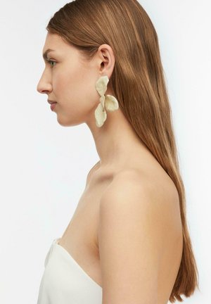 Profil kobiety z długimi, prostymi włosami, noszącej duże, abstrakcyjne kremowe kolczyki i biały, bezramiączkowy top na jednolitym tle.