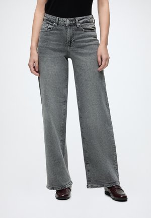 Person iført grå, høj talje, vidde jeans med forlommer og mørkebrune sko, stående mod en ensfarvet baggrund.
