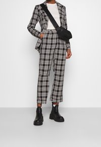 Osoba oblečená v černobílé kostičkované sako s zkrácenými kalhotami, bílou košili, černou přes rameno taškou a černými platformovými botami.