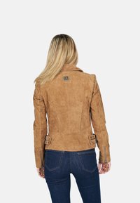 Bruine suède jas met een getailleerd ontwerp, benadrukt door gespspecificaties op de taille en ritsen op de mouwen. Eenvoudig logo op de achterkant.