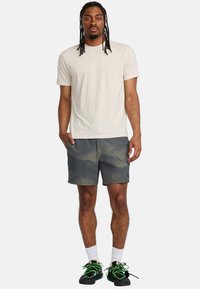 Beige Rundhals-T-Shirt, graue Camouflage-Shorts und schwarze Schuhe mit leuchtend grünen Schnürsenkeln. Das Modell steht mit einer Hand in der Tasche und präsentiert einen lässigen Stil.