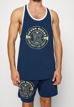 Mann trägt blaues ärmelloses Tanktop und Shorts-Set mit gelbem rundem "Seven Bucks" Logo und Text "Grind Gratitude Growth" auf der Vorderseite.