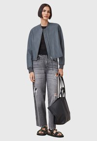 Giubbotto bomber in pelle grigia con zip frontale, abbinato a jeans grigi strappati. Una borsa tote nera e sandali flat completano l'outfit.
