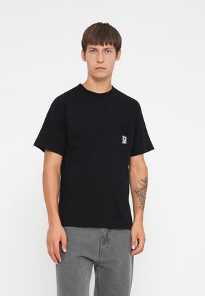 Black cotton t-shirt with a round neck, short sleeves, and a left chest pocket featuring a graphic design. Worn with gray jeans.