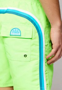 Costume da bagno verde neon con strisce laterali blu e bianche, vita elasticizzata e una tasca posteriore con una toppa blu con il logo "SUNDEK".