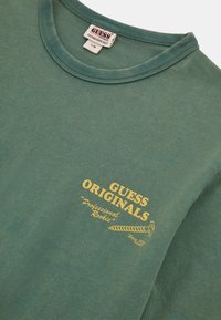 Grüner Baumwoll-Sweatshirt mit Rundhalsausschnitt, ausgestattet mit einem gelben Grafikdruck, auf dem "GUESS ORIGINALS" und "Professional Rookie" steht.