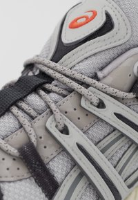 Harmaa urheilukenkä, jossa on teksturoitu kangasosa, harmaita ja mustia muovivaihtoehtoja sekä monimutkainen nauhadetaili, jossa on pieni oranssi logo.