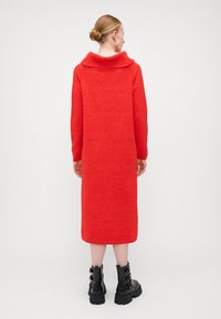 Abito rosso lavorato a maglia con un ampio colletto, maniche lunghe e lunghezza sotto il ginocchio. Indossato con stivali neri stringati per un contrasto.