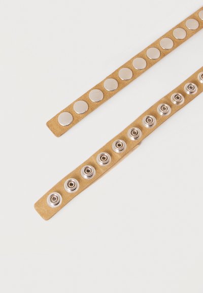 Deux lanières en daim beige, chacune avec une rangée de boutons-pression argentés, l’une montrant le dessus lisse et l’autre le côté à douille.