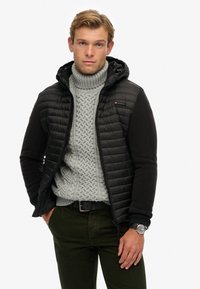 Schwarze gepolsterte Jacke mit Kapuze und Reißverschluss, kombiniert mit einem hellgrauen, grob gestrickten Pullover, grünen Hosen und einer Uhr. Glatter Stoff und strukturierte Form.
