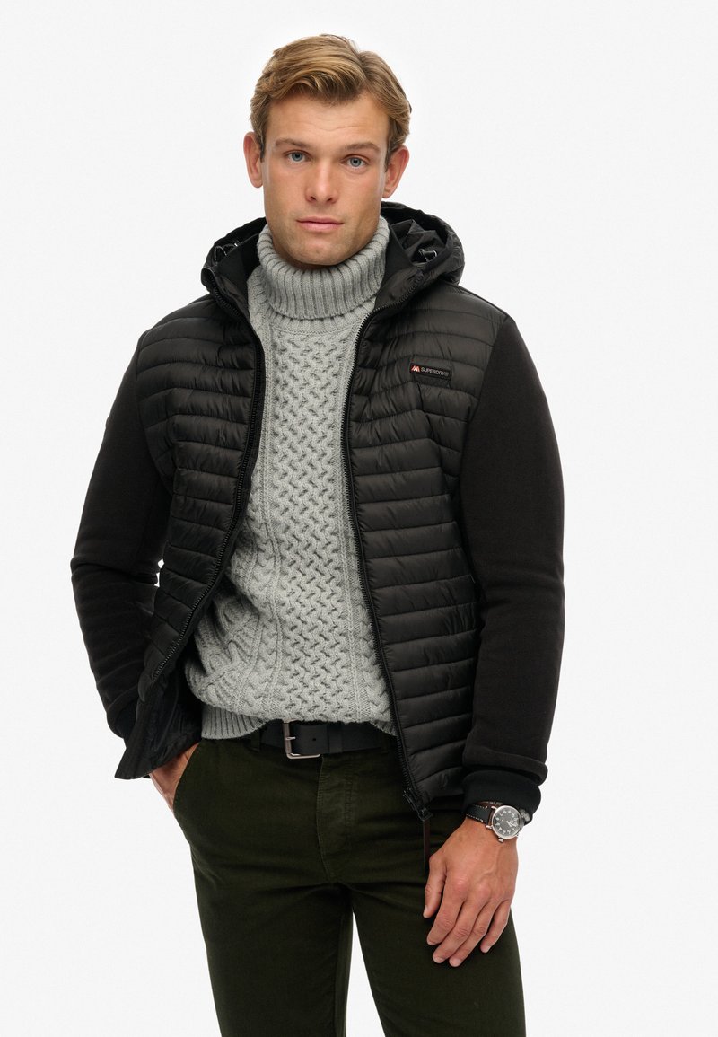 Schwarze gepolsterte Jacke mit Kapuze und Reißverschluss, kombiniert mit einem hellgrauen, grob gestrickten Pullover, grünen Hosen und einer Uhr. Glatter Stoff und strukturierte Form.