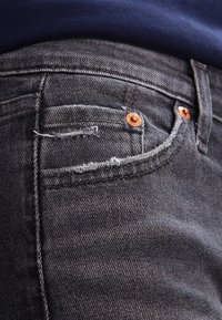 Los jeans de mezclilla gris oscuro presentan una textura desteñida, detalles de hardware en cobre y un pequeño bolsillo delantero. La tela muestra un deshilachado mínimo.