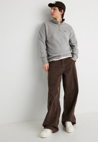 Νέος άνδρας με γκρι φούτερ με φερμουάρ μέχρι τη μέση, καφέ παντελόνια φαρδιάς γραμμής, λευκά αθλητικά παπούτσια και σκουρόχρωμο καπέλο, στέκεται μπροστά σε λευκό απλό τοίχο.