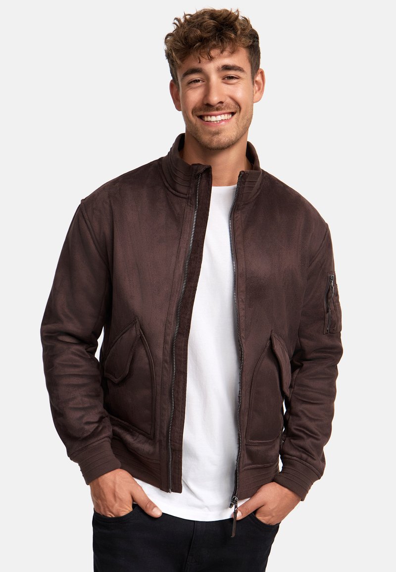 Cazadora bomber marrón con una textura suave, que presenta un cierre de cremallera, cuello acanalado y dos bolsillos frontales. Llevada sobre una camiseta blanca.