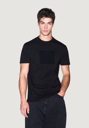 Młody mężczyzna z kręconymi ciemnymi włosami, ubrany w czarną koszulkę Armani Exchange i ciemne dżinsy, stoi z jedną ręką w kieszeni.