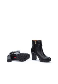 Botas negras de cuero hasta el tobillo con tacones gruesos, cremallera lateral, detalle de hebilla y suelas de goma texturizadas que presentan un patrón de tracción.