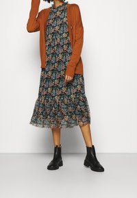 Lang blomsterkjole i mørkt stof med flæser nederst, kombineret med en orange cardigan og sorte ankelstøvler.