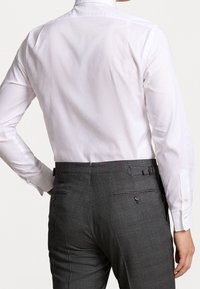 Biała koszula z długim rękawem o gładkiej fakturze i klasycznym kołnierzyku, zestawiona z szarymi spodniami z delikatnym wzorem i detalami kieszeni tylnej.