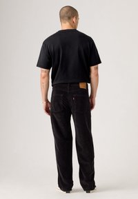 Homme avec une coupe très courte, portant un t-shirt noir, un pantalon en velours côtelé noir et des baskets blanches, debout dos à un fond uni.