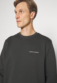 Felpa grigia scura con scollatura a girocollo, caratterizzata da un piccolo logo ricamato bianco. Realizzata in un tessuto liscio e strutturato.