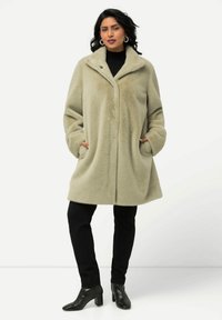 Cappotto in pelliccia sintetica verde chiaro con ampio colletto, tasche laterali e chiusura con un solo bottone, abbinato a pantaloni neri e stivaletti alla caviglia.