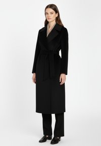 Vrouw in een lange zwarte mantel met riem over zwarte broeken en zwarte instappers, staand tegen een effen witte achtergrond.