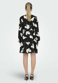 Robe noire avec un motif floral blanc, manches longues, taille cintrée et jupe évasée. Portée avec des talons aiguilles noirs, vue de dos.