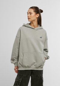 Lys grå oversized hættebluse lavet af blødt stof, med en frontlomme og et lille logoaccent på brystet. Justerbar hætte.