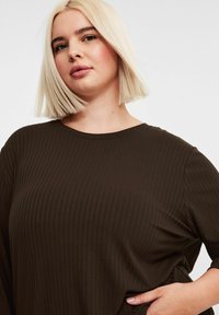 Blondin nainen, jolla on suora bob-leikkaus, pukeutuneena tummanruskeaan ribbipaitaan, käsi vyötäröllä, neutraali ilme, valkoinen tausta.