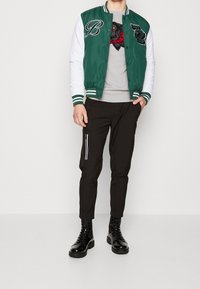 Man med en grön och vit varsityjacka, grå tröja med en vargdesign, svarta byxor med dragkedjeficka och svarta snörstövlar.