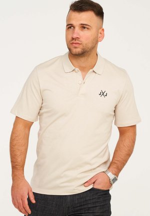 Beige Poloshirt mit kurzen Ärmeln, zwei Knöpfen und einem kleinen schwarzen Logo auf der linken Brust. Wird mit dunklen Jeans getragen.