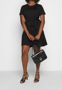 Robe noire à manches courtes avec un détail de taille nouée, associée à un sac à main en cuir crocodile noir et des talons noirs à lanières.