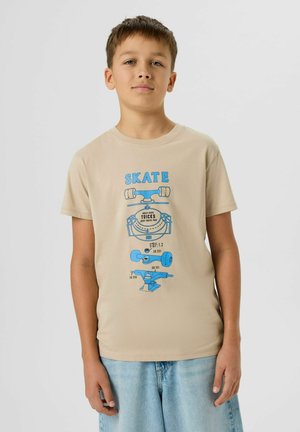 Ragazzo che indossa una maglietta beige con illustrazione di pezzi di skateboard blu e pantaloncini di jeans azzurri, in piedi davanti a uno sfondo grigio uniforme.