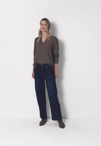 Kobieta stoi przy białej ścianie, ubrana w brązową bluzkę z długim rękawem, ciemne szerokie dżinsy oraz brązowe botki za kostkę.