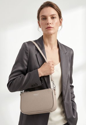 Femme portant un blazer gris et un haut crème tenant un sac à bandoulière beige avec un logo sur la sangle avant sur fond blanc.