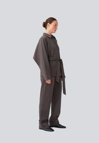 Giacca grigia su misura con ampio collo e chiusura a bottoni, abbinata a pantaloni larghi coordinati, fissati con una cintura di tessuto e texture sottili.